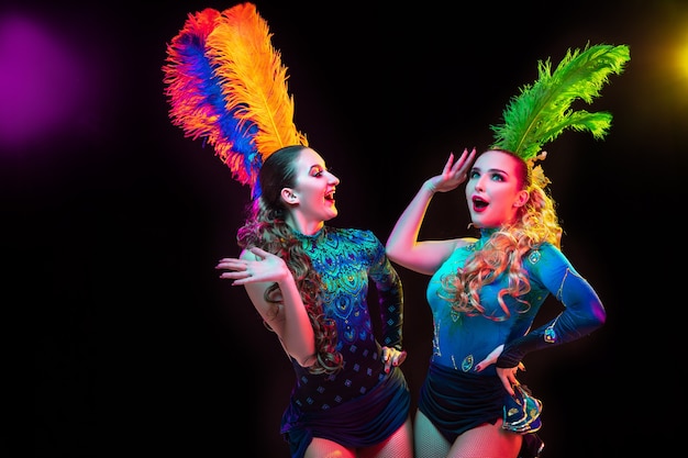 Beautiful young women in carnival, stylish masquerade costume with feathers on black wall in neon light