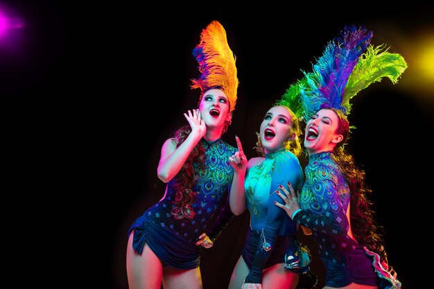 Beautiful young women in carnival, stylish masquerade costume with feathers on black wall in neon light