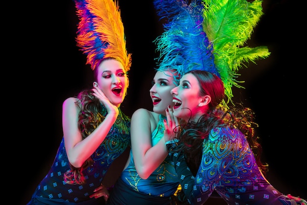 Beautiful young women in carnival, stylish masquerade costume with feathers on black wall in neon light