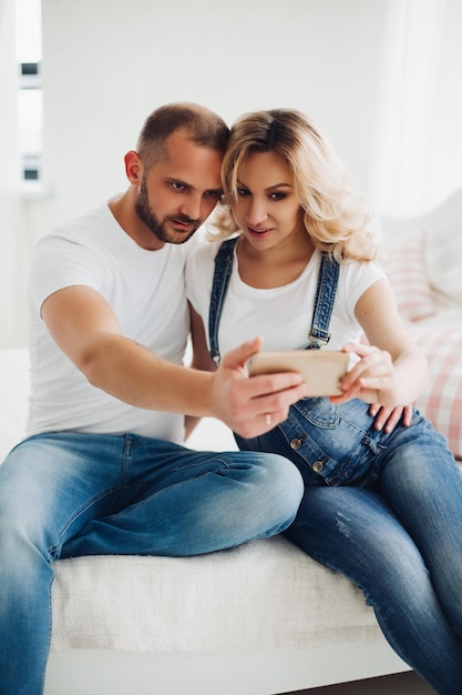 Beautiiful couple of pregnant woman and her husband wearing in jeans sitting kissing each other and taking self portrait Young mom and dad holding smart phone and taking photo