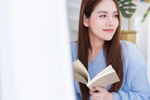 Красавица азиатская женщина в белом платье с длинными волосами читает книгу на белой кровати с моллюском и веселым белым фоном интерьера спальни
