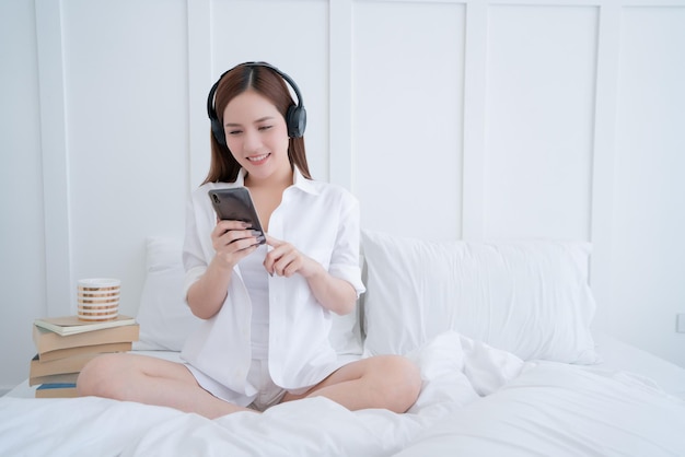 아름다운 아시아 여성 흰색 드레스 긴 머리는 헤드폰을 끼고 조개와 밝은 흰색 침실 인테리어 배경으로 흰색 침대에서 유형 검색 스마트폰을 사용합니다.