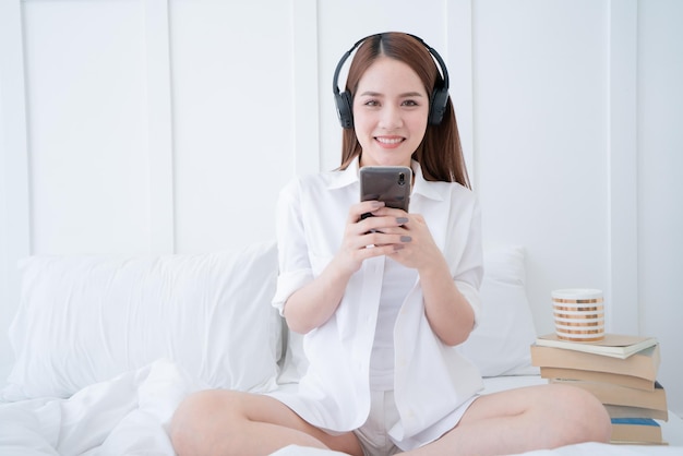 Beauty asian female white dress long hair wear headphone and use type search smartphone on white bed with clam and cheerful white bedroom interior background