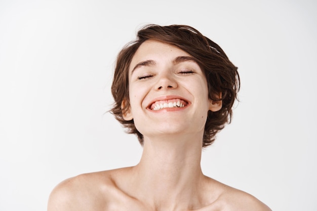 Beauty. Close-up of beautiful and natural girl with white smile and naked shoulders laughing, smiling carefree. Young woman without makeup showing effect after cosmetics, white wall