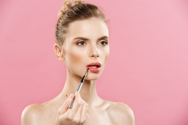 Beauty Concept - Woman applying red lipstick with pink studio background. Beautiful girl makes makeup.