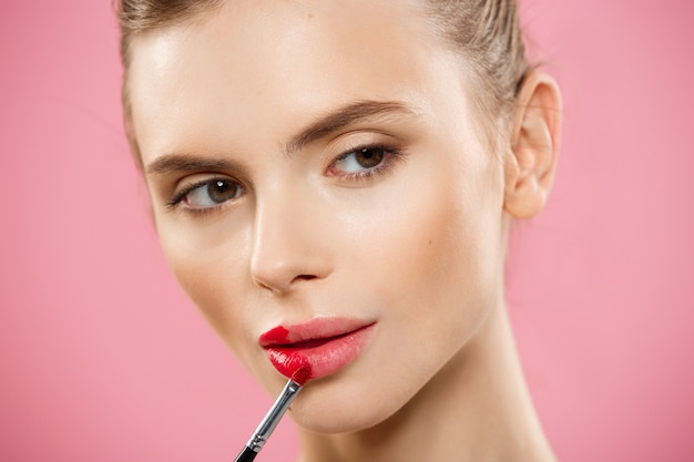 美容の概念 - ピンクのスタジオの背景と赤い口紅を適用している女性。美しい女の子が化粧をする。