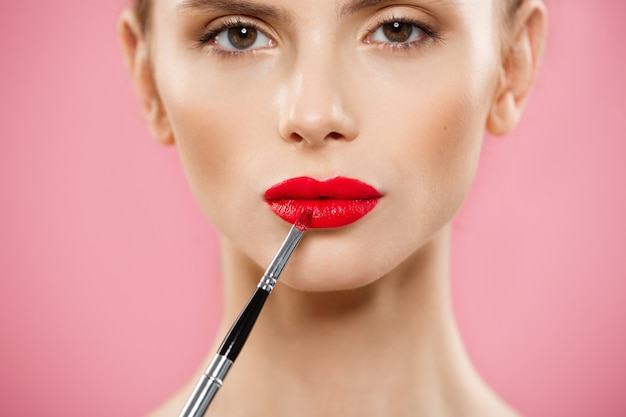 Free photo beauty concept - woman applying red lipstick with pink studio background. beautiful girl makes makeup.