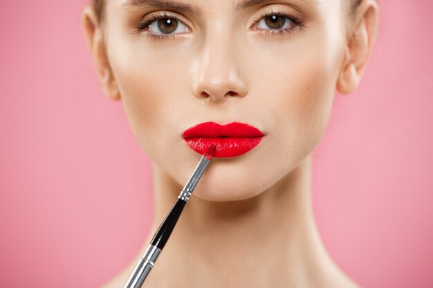 Beauty Concept - Woman applying red lipstick with pink studio background. Beautiful girl makes makeup.