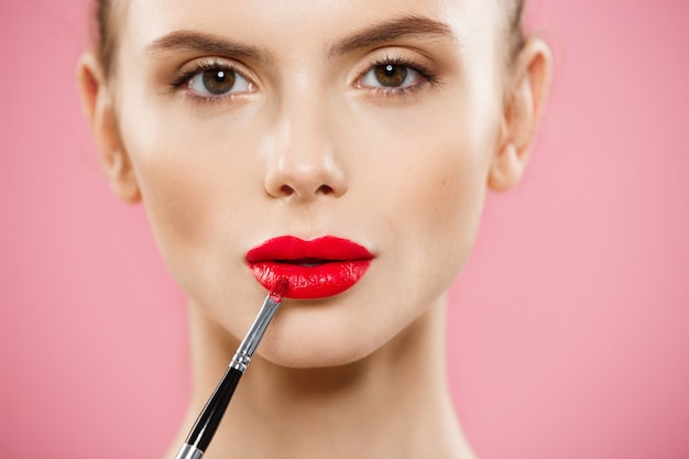 Beauty Concept - Woman applying red lipstick with pink studio background. Beautiful girl makes makeup.