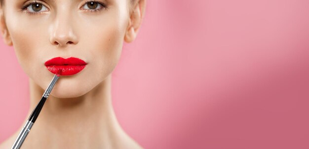 Beauty Concept Woman applying red lipstick with pink studio background Beautiful girl makes makeup