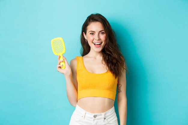 Beauty and cosmetics. Smiling woman with glowing curly hairstyle, showing brush without hair strands, taking care or health, standing over blue background.