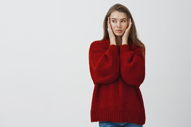 Beauty, cosmetology and lifestyle concept. Attractive stylish and sassy adult female model in loose red stylish sweater touching skin looking left, enjoying smooth skin after applying mask