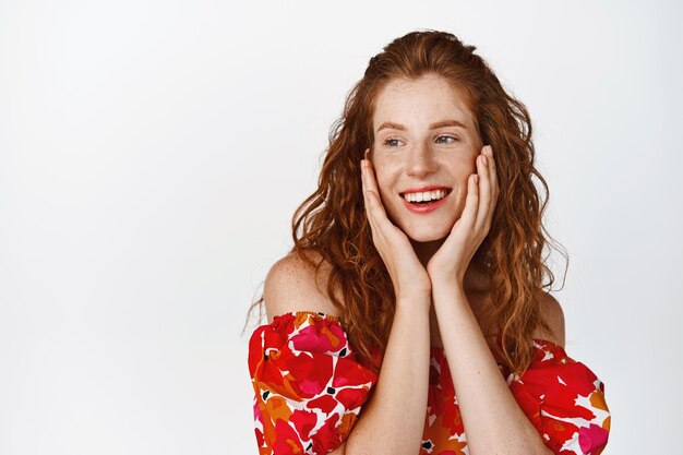 Free photo beauty cute smiling redhead woman in summer floral dress gazing flirty aside and grinning standing happy against white background