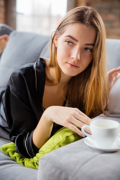 Beauty Day for yourself. Woman wearing silk robe doing her daily skincare routine at home. Lying on sofa, reading magazin, drinking coffee. Concept of beauty, self-care, cosmetics, youth. Close up.