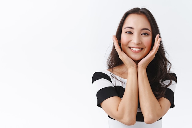 Beauty, emotions and cosmetology concept. Attractive, beautiful young asian girl touching pure, clean no blemishes skin and smiling, looking lovely and deligted, standing silly white background