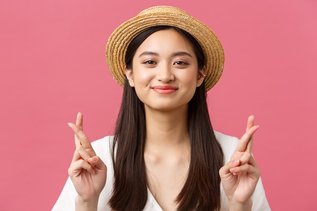 Beauty, emotions and leisure and vacation concept. Close-up of hopeful dreamy asian girl wishing for summer trip to sea, wearing straw hat, cross fingers good luck and smiling optimistic.