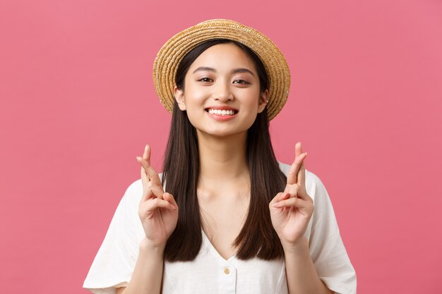 Beauty, emotions and leisure and vacation concept. Close-up of hopeful dreamy asian girl wishing for summer trip to sea, wearing straw hat, cross fingers good luck and smiling optimistic.