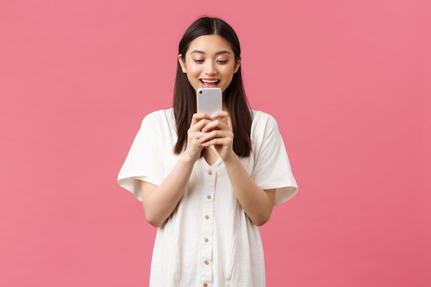 Концепция красоты, эмоций и технологий. Взволнованная азиатская девушка-блоггер смотрит на мобильный телефон с удивленным счастливым лицом, отправляет текстовые сообщения или использует приложение, смотрит видео на смартфоне, розовый фон.