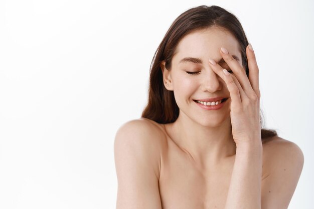 Beauty face. Smiling caucasian woman touching healthy skin portrait. Beautiful happy girl model with fresh glowing hydrated facial skin and natural makeup on blue background at studio