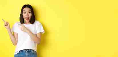 Free photo beauty and fashion concept beautiful asian woman in white tshirt pointing fingers left standing over