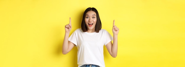 Free photo beauty and fashion concept beautiful asian woman in white tshirt pointing fingers up standing over y