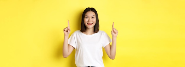 Free photo beauty and fashion concept beautiful asian woman in white tshirt pointing fingers up standing over y