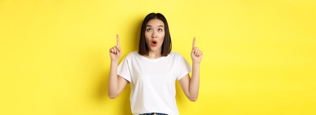 Free photo beauty and fashion concept beautiful asian woman in white tshirt pointing fingers up standing over y