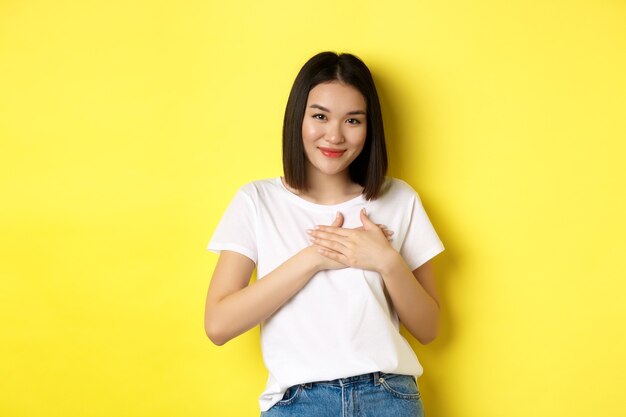 Beauty and fashion concept. Heartfelt asian girl holding hands on heart and smiling touched, thanking you, standing over yellow background