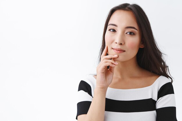 Beauty fashion and healthcare concept Tender feminine young asian woman with clean skin touching cheek and jawline and smiling misterious gazing camera pensive standing thoughtful