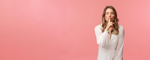 Beauty fashion and women concept Portrait of feminine and flirty caucasian woman hiding secret asking keep silent shushing with sensual coquettish smile wear white dress pink background