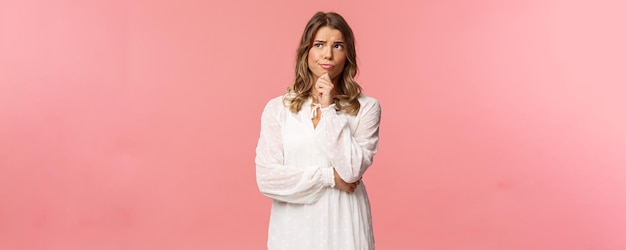 Beauty fashion and women concept Portrait of uncertain seriouslooking feminine young blond woman in white dress touch chin smirk and frowning thoughtful look away thinking