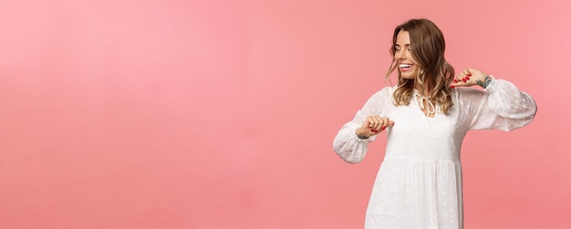 Foto gratuita bellezza moda e concetto di donne ragazza carina tenera e spensierata che si gode il tempo primaverile indossando un abito bianco che balla e distoglie lo sguardo con un sorriso raggiante divertendosi sullo sfondo rosa