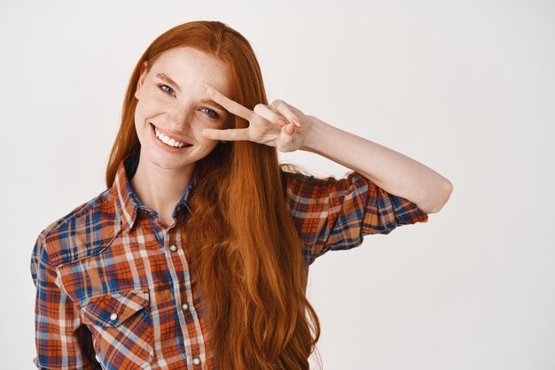 Beauty Image of young woman with long healthy red hair pale skin and blue eyes smiling happy and showing peace sign standing over white background
