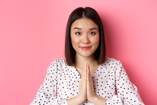 Free photo beauty and lifestyle concept. close-up of cute asian woman smiling, showing thank you gesture, holding hands in pray, standing over pink background