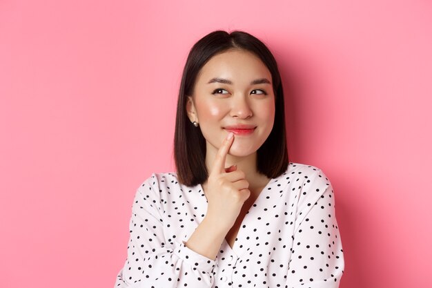 Beauty and lifestyle concept. Close-up of dreamy asian girl smiling, looking left and thinking, making choice, standing over pink background