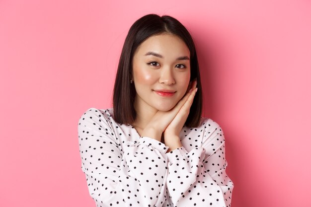 Beauty and lifestyle concept. Close-up of pretty korean girl leaning on palms, looking at camera with admiration, standing over pink background.