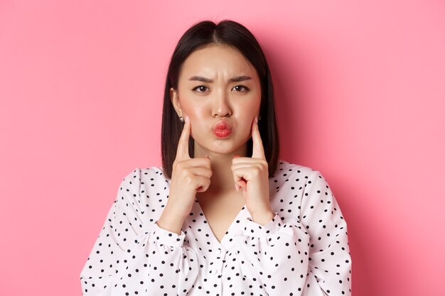 Beauty and lifestyle concept. Cute asian woman making grumpy face, poking cheeks and grimacing, standing over pink background.