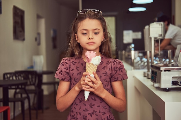ファッショナブルなドレスを着た美女が、アイスクリームパーラーに立ってストロベリーアイスクリームを持っています。