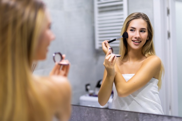 Beauty, make up, cosmetics, morning and people concept - young woman with makeup brush and contouring and highlighting palette looking to mirror at home bathroom