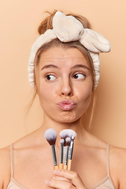 Foto gratuita concetto di bellezza e trucco. la giovane e graziosa donna premurosa con i capelli pettinati mantiene le labbra arrotondate tiene i pennelli cosmetici pensa a cosa guardare per la data che avrà la procedura di applicazione dei cosmetici