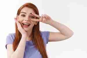 Free photo beauty makeup and tenderness concept cheerful goodlooking caucasian redhead female in purple tshirt touching pure clean skin no blemishes show peace gesture over eye smiling happy