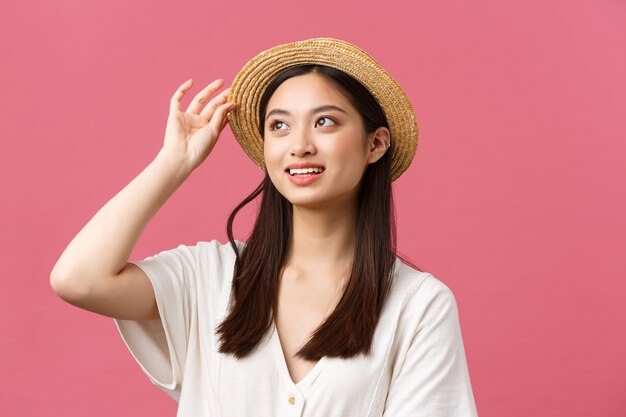Красота, эмоции людей и концепция досуга и отдыха. Прекрасная азиатская женщина делает покупки в магазине, выбирает новую соломенную шляпу, радостно улыбается, покупает летнюю одежду на розовом фоне.