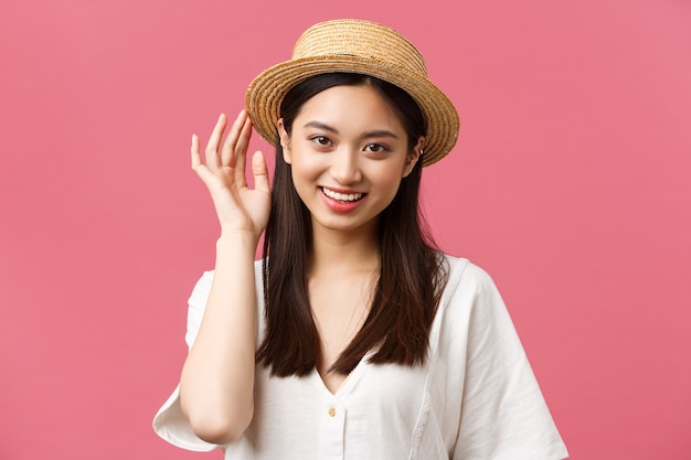Beauty, people emotions and leisure and vacation concept. Lovely asian woman shopping in store, picking new straw hat, smiling delighted, buying summer outfit over pink background.