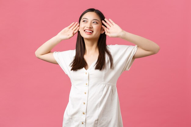 Beauty, people emotions and summer leisure concept. Dreamy and happy korean girl with perfect skin, wearing dress, looking at sun on vacation, tourist enjoying perfect day-off, pink background.