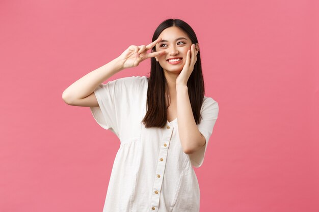 Beauty, people emotions and summer leisure concept. Dreamy and romantic cute smiling asian girl looking away thoughtful while touching soft skin on face and show peace sign, pink background