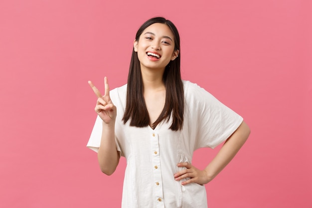 Beauty, people emotions and summer leisure concept. Enthusiastic happy japanese girl laughing and smiling, showing kawaii peace sign in white cute dress, pink background