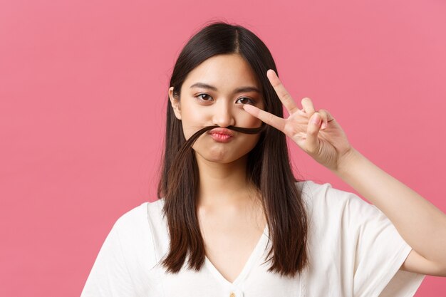 Beauty, people emotions and summer leisure concept. Playful and silly asian girl fool around, showing peace sign and hold hair strand over lip like moustache, pink background.