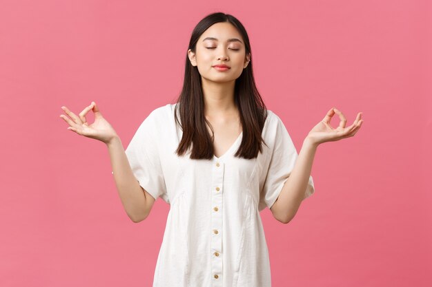 Beauty, people emotions and summer leisure concept. Relaxed and calm, determined young asian woman meditating in white dress, close eyes and do yoga breathing exercises, pink background.