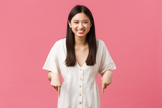 Beauty, people emotions and summer leisure and vacation concept. Carefree outgoing cute asian girl in white dress laughing from joy, pointing fingers down, inviting to party or event.