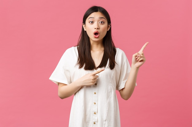 Beauty, people emotions and summer leisure and vacation concept. Impressed cute asian girl pointing fingers upper right corner with dropped jaw and surprised face, stand pink background